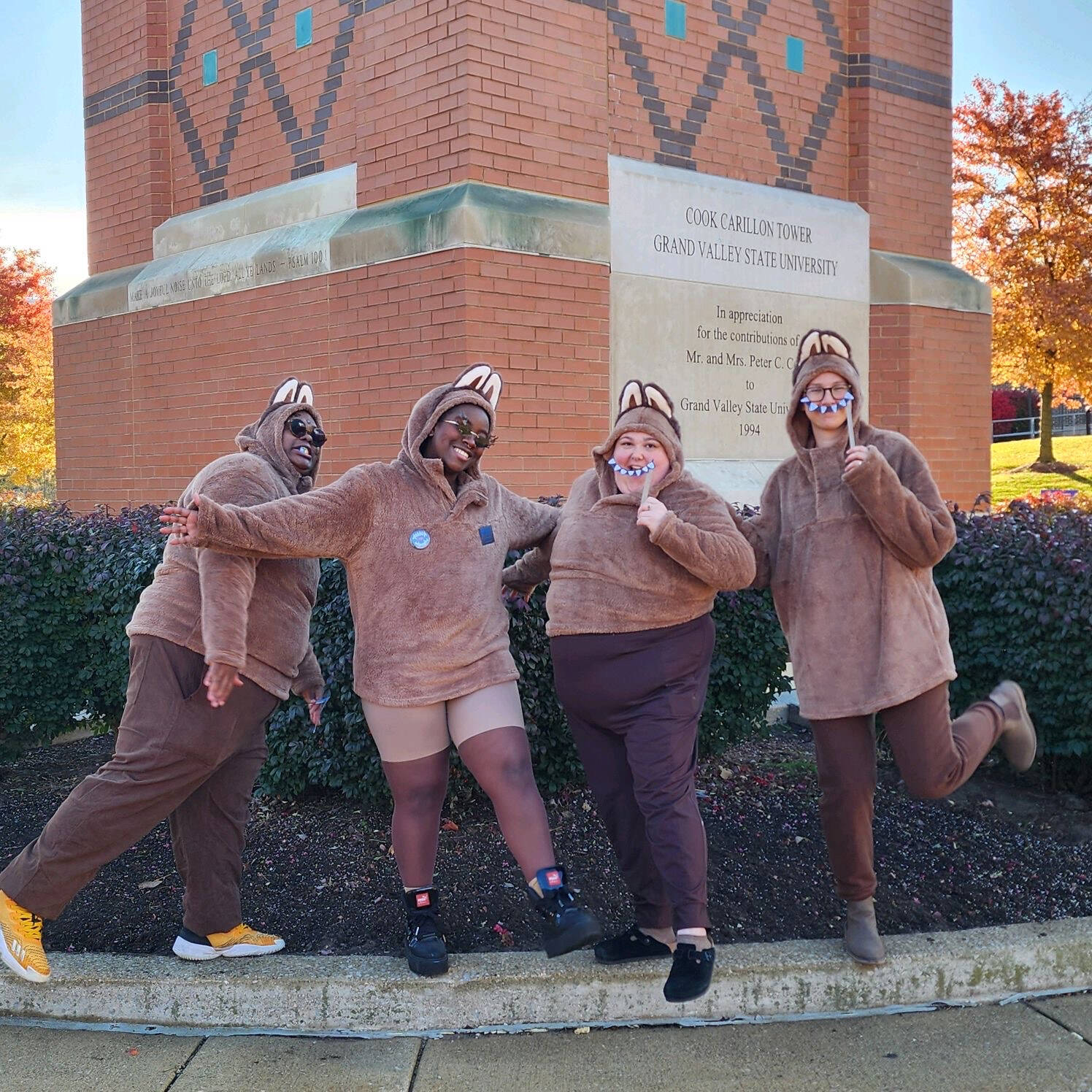 Four people dressed in fuzzy brown Labubu-inspired costumes with oversized ears and toothy smiles pose together outdoors in front of a brick building. One person extends their arms playfully while the others smile for the camera.